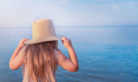 Child girl holding hat with hands and looking into the seaの写真素材