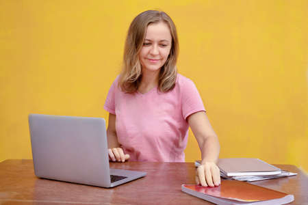 Woman sitting at work table with laptop and reaching for book on yellow background