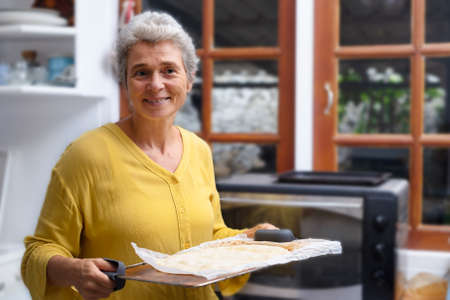 Portrait of an elderly woman with pastries in the kitchen. Retiree preparing food for the familyの写真素材