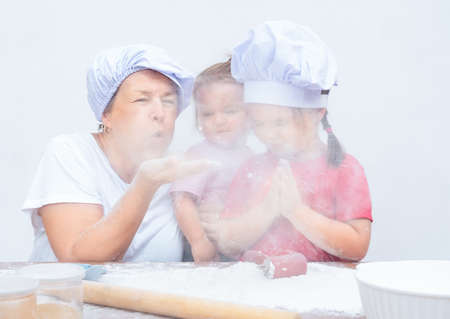 Grandmother and grandchildren blow on flour. Woman with children preparing pastries togetherの写真素材