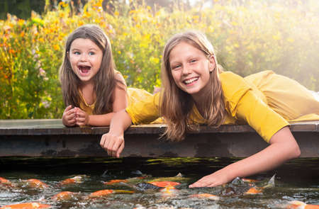 Girl sisters lie on the bridge and feed the fish in the park outdoorsの写真素材