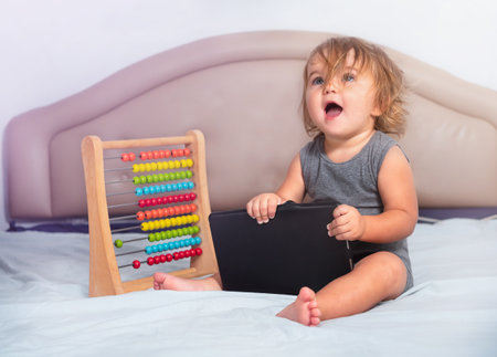 The baby sits on the bed with a tablet and bills. Skills developmentの写真素材