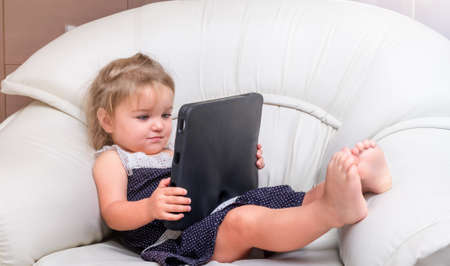 Child girl lies in the chair and plays on the tablet