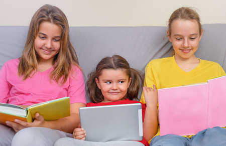 A child girl with a tablet looks into the books to a teenager on the couchの写真素材