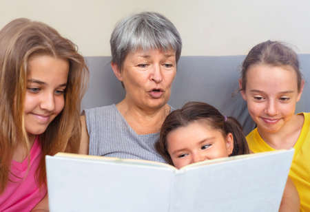Grandmother reads a book with stories to her granddaughters sitting together on the couchの写真素材
