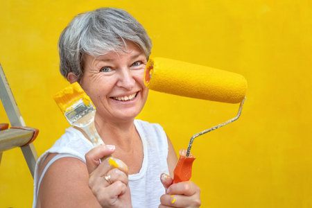 Woman sitting on a stepladder with a brush and a paint roll near a yellow wallの写真素材