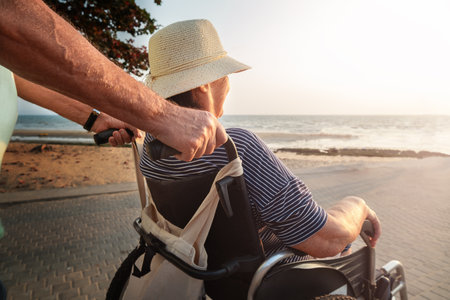 Senior husband pushing senior wife in wheelchair along sea lineの写真素材