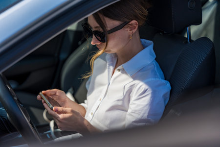 A woman in sunglasses and a white shirt sits in a car, holding a smartphone tilted horizontally.