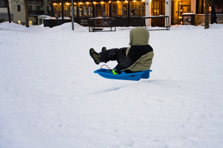 A child in a green and black winter jacket sleds down a snowy hill on a blue plastic sled.の写真素材
