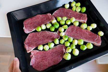 Raw pork slices and Brussels sprouts on black oven tray, healthy meal preparationの写真素材