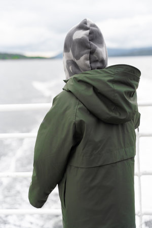 Child in hooded jacket watching sea wake from ferry deck in Norway in the summer.