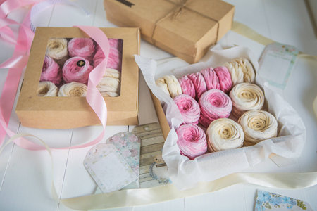 Delicate cream and pink marshmallows, packed in boxes of kraft paper with satin ribbons on a white wooden table.の写真素材