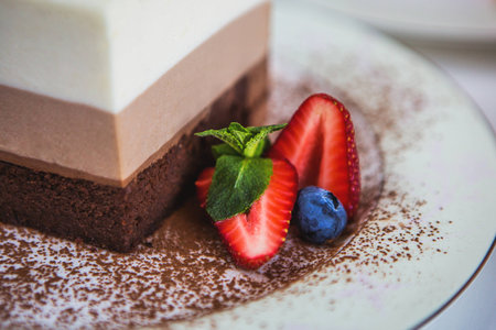 A piece of a mousse cake "three chocolate" stands on a plate, sprinkled with grated chocolate, and decorated with berries and biscuits. A plate with a cake stands on a white wooden table.の写真素材