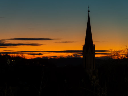 A church spire stands tall against a stunning sunset, with vibrant orange and blue hues illuminating the sky. The silhouettes of trees add depth to the evening scene.の写真素材