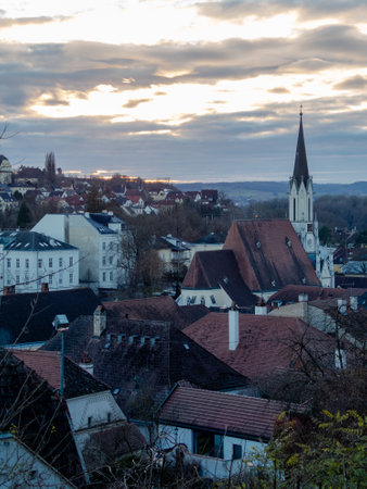 Rooftops and historic buildings stretch across the landscape as the sun sets, casting a warm glow. A tall church steeple rises above the town, enhancing the picturesque view.の写真素材