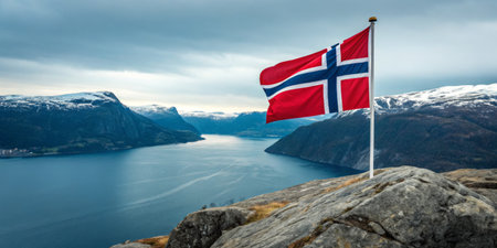 A vibrant Norwegian flag flutters proudly on a rocky ledge, overlooking a calm fjord surrounded by majestic mountains. The cloudy sky adds a dramatic ambiance to the breathtaking view.の素材