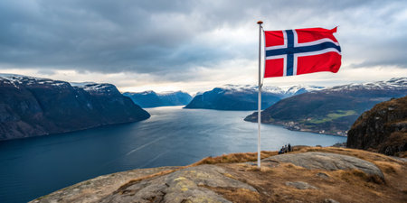 A breathtaking fjord stretches into the distance, framed by steep mountains and a cloudy sky, with a Norwegian flag flying proudly on a rocky outcrop.の素材