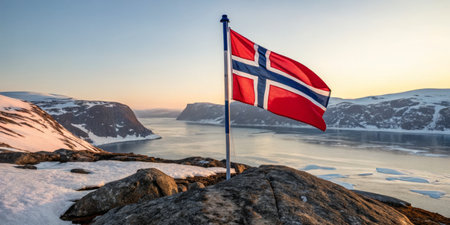 A stunning view of the Norwegian flag proudly flying atop a rocky outcrop as the sun sets over a serene fjord, surrounded by icy mountains and calm waters.の素材