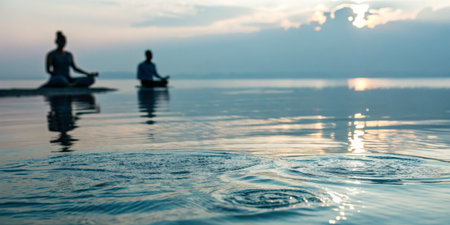 Two individuals sit cross-legged on the shore, meditating peacefully as the sun rises. The tranquil...