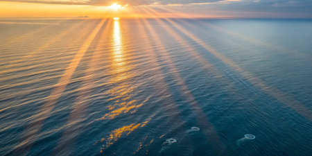 Golden sunlight radiates over tranquil sea waters at sunset, casting shadows and creating a serene atmosphere. Gentle waves reflect the vibrant colors in the sky.の写真素材