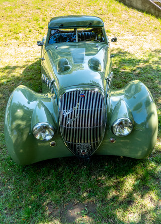 A striking vintage car with a glossy green exterior is prominently showcased on the grass. Sunlight...