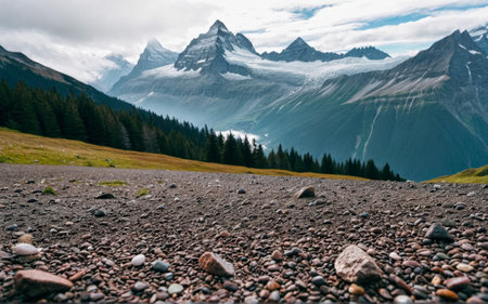 Majestic mountain range towers over a green valley, with rocky terrain in the foreground. Clouds...