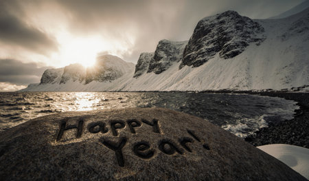 A rocky shore reflects the warm glow of the setting sun as snow-capped mountains rise in the distance. The words Happy Year! are carved into a stone, welcoming the new beginning.の写真素材