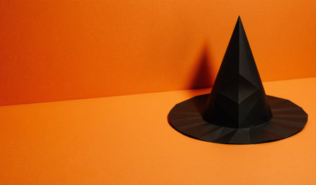 A striking black paper witch hat sits alone on a bright orange surface. This eye-catching display is perfect for Halloween events, themed parties, or artistic projects celebrating spooky vibes.の写真素材