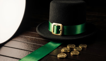 A festive black hat adorned with a green ribbon sits on a wooden surface, accompanied by sparkling gold coins. Perfect for St. Patrick's Day celebrations, it radiates luck and joy.の写真素材