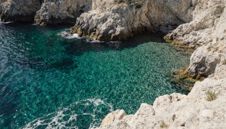 Sunlight dances on the surface of calm turquoise waters, reflecting against rocky cliffs. Gentle waves lap at the shore, creating a sense of peaceful tranquility by the sea.の写真素材