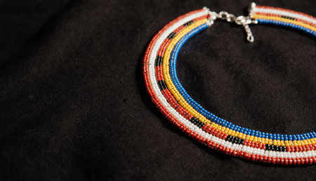 A beautifully crafted beaded necklace lies on a dark fabric surface. Its vibrant colors of red, blue, yellow, and black create intricate patterns, reflecting cultural artistry and tradition.の写真素材