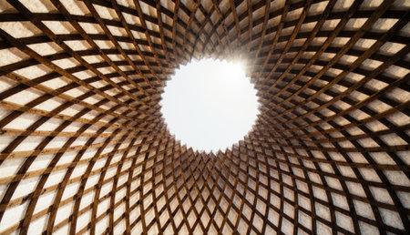 Looking upward through an intricate wooden dome, sunlight spills through a central opening, illuminating the unique geometric patterns. This captivating structure invites awe and inspiration.の写真素材