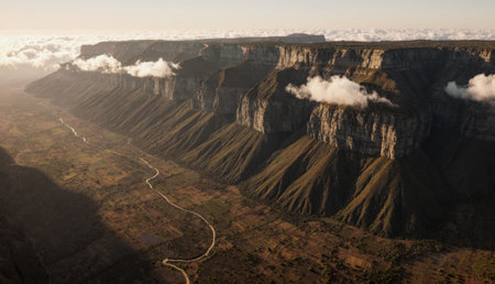 Stunning cliffs rise dramatically over a lush valley, with a gentle river meandering below. Soft, fluffy clouds drift across a golden sky as daylight fades into evening.の写真素材