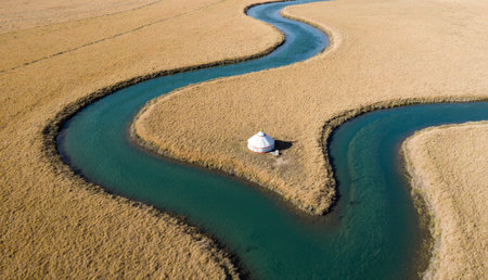A serene river winds gracefully through golden grasslands, showcasing a traditional yurt perched on the bank. The sky is bright and clear, highlighting the peacefulness of the scene.の写真素材