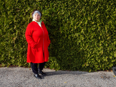 A woman stands proudly wearing a bright red coat, contrasting beautifully with a dense green hedge. The sun casts a warm glow, highlighting the serene atmosphere of the autumn day.の写真素材