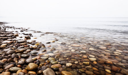 Soft waves gently kiss the smooth stones scattered along the shoreline. A peaceful atmosphere envelops the scene as dusk settles over the calm waters, creating a serene retreat.の写真素材