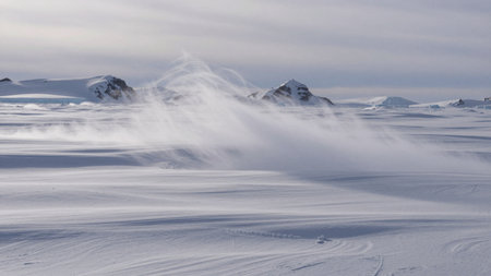 Strong winds move snow across the flat ice landscape. Mountains are visible in the distance under a cloudy sky. The scene shows the effects of weather in a cold environment.の写真素材