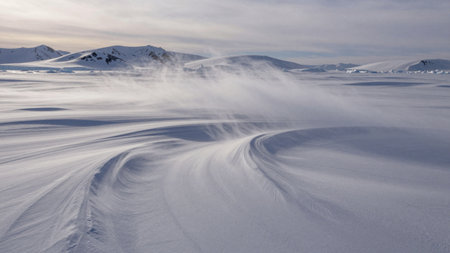 Wind blows snow across a flat frozen landscape showing tire tracks. Mountains are visible in the distance under a cloudy sky. This scene captures the harsh beauty of winter.の写真素材