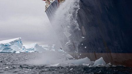 A large ship cuts through icy waters in Antarctica, causing chunks of ice to break and splash. The scene shows surrounding icebergs and cloudy skies, creating a dramatic landscape.の写真素材