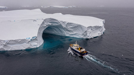 A boat moves through icy waters close to a large glacier. The setting shows dark water and a prominent ice formation. The scene captures a rugged and cold environment.の写真素材