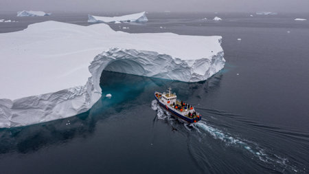 A boat moves through dark waters surrounded by large icebergs. The sky is overcast, and ice formations are visible in the distance. People are onboard enjoying the view.の写真素材