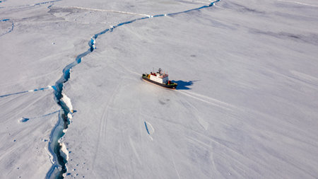 A boat navigates through frozen water in the Arctic. The scene shows ice cracks and clear daylight. The remote area highlights the beauty of the landscape and the challenges of travel.の写真素材