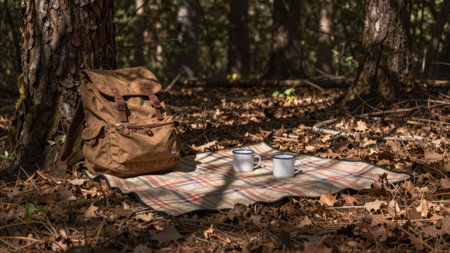Two mugs of steaming coffee rest on a soft blanket spread on the forest floor adorned with dried...