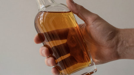 A hand gently grips a clear glass bottle filled with a warm amber liquid. The soft light highlights the contours and textures, creating a serene moment of contemplation.の写真素材