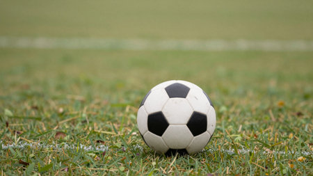 A classic black and white soccer ball sits on lush green grass beside a soccer field. The bright...