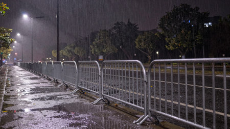Taken at night, depicting a wet, asphalt road with a row of metal barricades on either side. The...