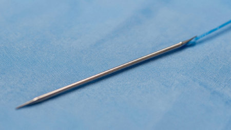 Of a needle lying on a light blue fabric surface. The needle is made of stainless steel and has a pointed tip with a smooth, polished finish. It is positioned in the center of the.