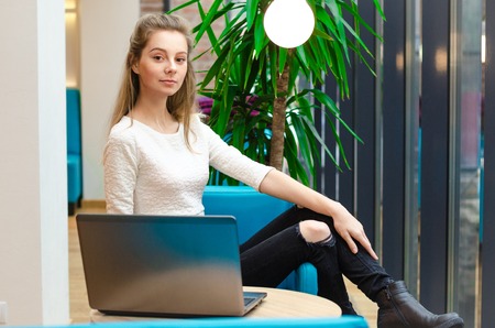 Portrait of beautiful smiling woman sitting on a comfortable chair in a cafe with black laptop. Pretty student doing homework with laptopの写真素材