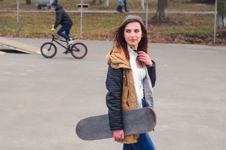 Portrait of of a young happy girl holding a skateboard. Skate park cyclist in the backgroundの写真素材