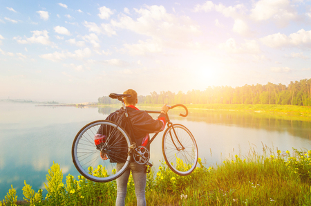 man stand with bicycle in hands on the hill and looking at the lake with azure water and sunriseの写真素材